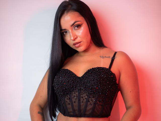 HikariChloe with long black hair, wearing a black beaded bra, exposing her tits, tattoo on right shoulder, red and white light on her face.