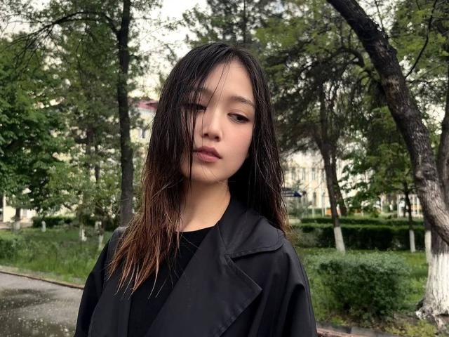 ShellaKircher mit nassen, schwarzen Haaren, die eine schwarze Jacke trägt und in einem Park steht, blickt leicht zur Seite und hat einen neutralen Gesichtsausdruck. Ihr Hintern ist nicht sichtbar..