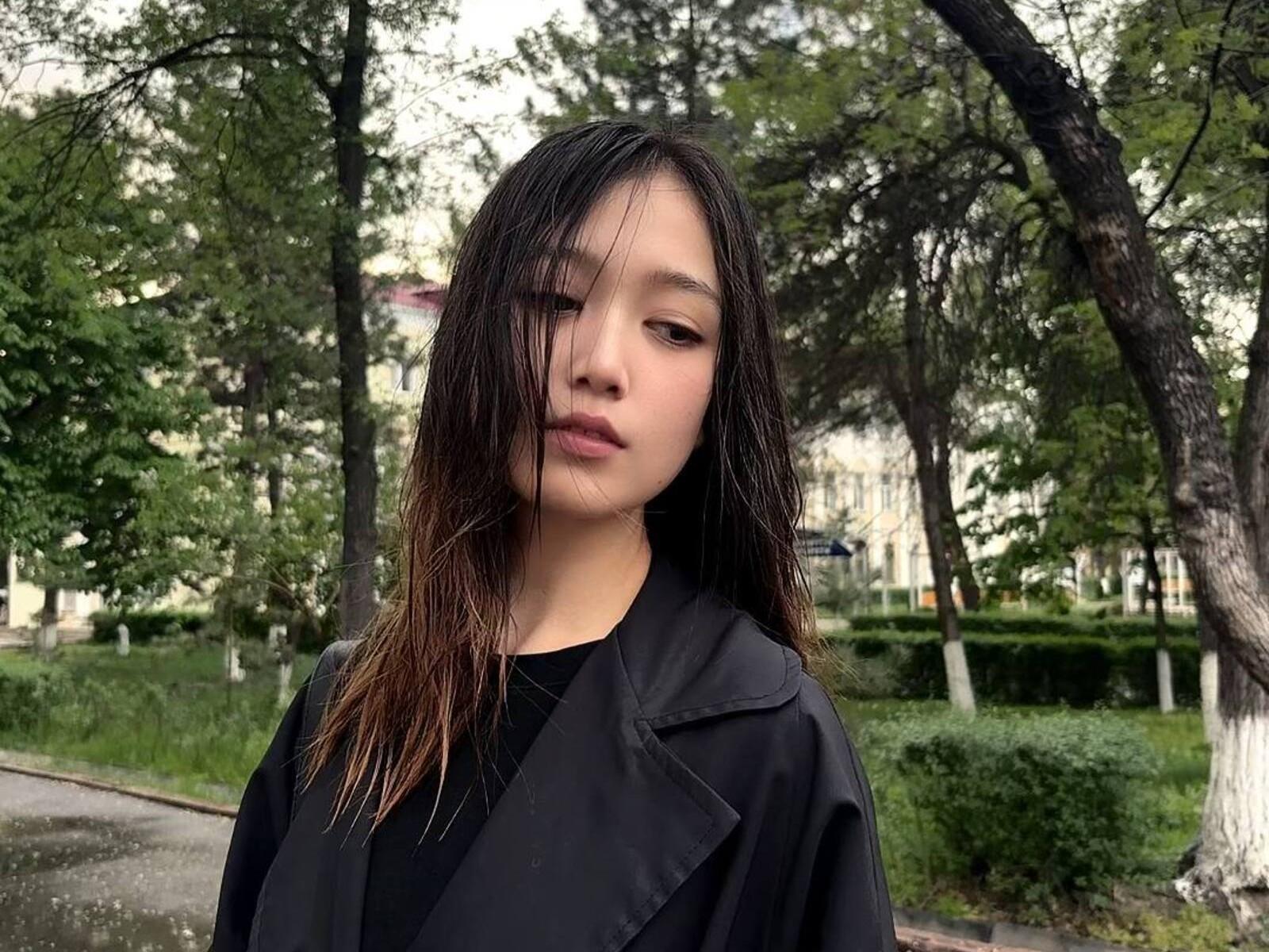 ShellaKircher with wet black hair, wearing a black jacket, standing in a park, looking slightly to the side with a neutral expression. Her ass is not visible.