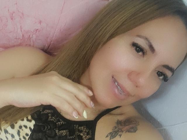 AmberDillucca with long brown hair, smiling, showing teeth, wearing black lace top, hand near chin, visible rose tattoo on shoulder, eyeliner, and pink nail polish.