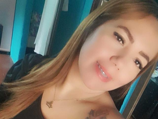 Amber Dillucca, con lunghi capelli castani, sorride mostrando labbra rosate, occhi marroni e un piccolo naso, indossa una maglietta nera e una collana con un ciondolo, e mostra un tatuaggio..