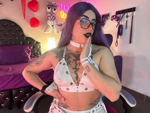 LilithDark69 with purple hair, glasses, and black lipstick, wearing a white, buttoned, cleavage-revealing top, showing tattoos, and a white choker, touching her lip with a finger.