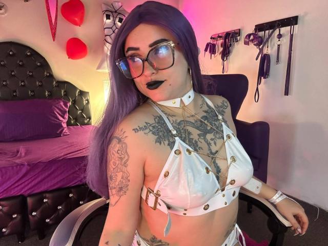 LilithDark69 with purple hair, glasses, black lipstick, white leather bra with gold studs, revealing tattoos, sitting in a chair, bedroom with purple bed, BDSM wall hooks in background.
