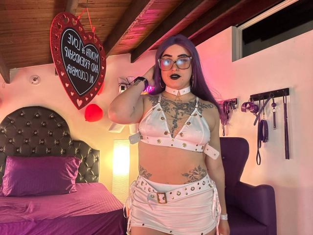 LilithDark69 with purple hair, glasses, black lipstick, white leather crop top and skirt, tattoos, holding hair, standing in bedroom with purple bed, white wall, and wooden ceiling.