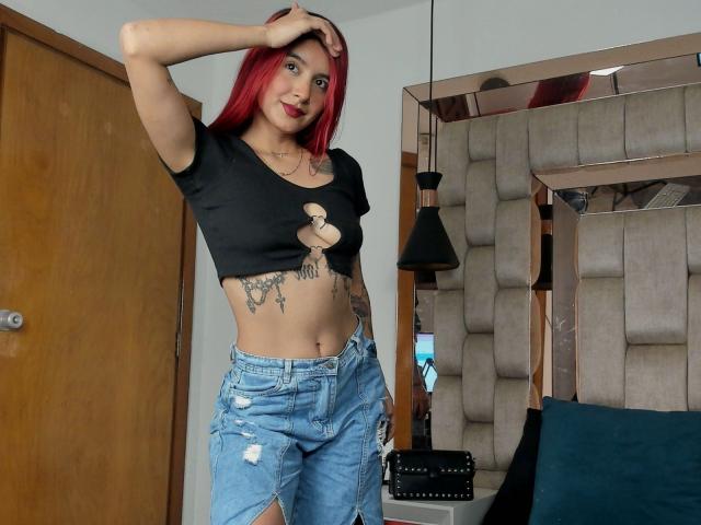 LeoFireX, con cabello rojo, una blusa negra con aberturas que muestra poco el busto y jeans azules rotos, tiene tatuajes en el abdomen y está de pie en una habitación, con una mano en la cabeza..