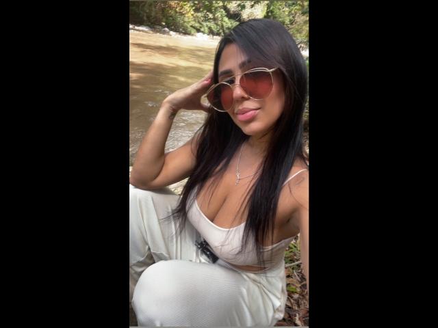 NahomyStone with long black hair, tan skin, wearing a white crop top and white pants, large brown sunglasses, and a necklace, sitting by a stream, smiling.
