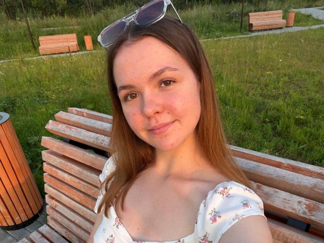 JeseniaPiontek has light skin, brown eyes, and straight brown hair. She's wearing a floral top, sitting on a wooden bench with sunglasses on her head, smiling.