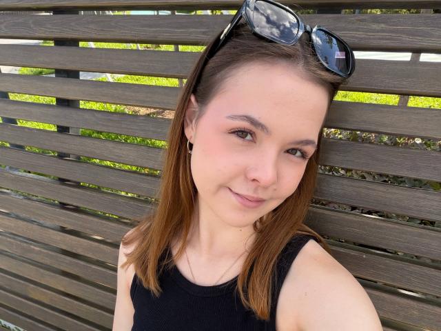 JeseniaPiontek with brown hair, black tank top, small hoop earrings, and sunglasses on her head, smiling softly at the camera, sitting on a wooden slat bench.