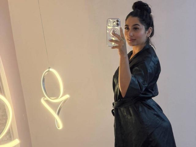 Jasmine, gekleed in een zwarte zijden kimono, staat met haar zij aan de camera, maakt een selfie met haar telefoon, waarbij haar achterkant en een zelfverzekerde glimlach te zien zijn, met donker haar in een knot..