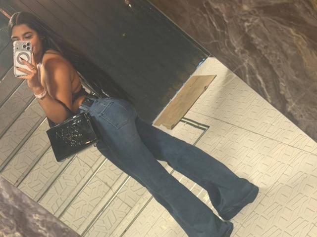 JasmineRodes indossa jeans a zampa d'elefante blu, mettendo in mostra i glutei, è senza top con lunghi capelli castani, tiene una borsa nera e sta scattando un selfie allo specchio con un iPhone..