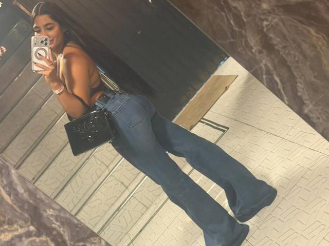 JasmineRodes, senza top, con jeans blu a zampa d'elefante, che tiene una borsa nera, sta scattando un selfie allo specchio, mostrando il sedere, con lunghi capelli scuri, che guarda indietro, con il telefono in mano..