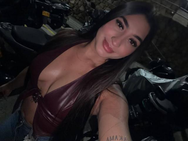 JasmineRodes with long dark hair, wearing a deep maroon leather top, showing her tits, smiling, taking a selfie in a dimly lit garage, with a motorcycle behind her.