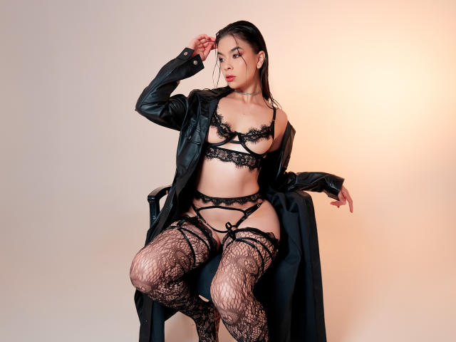 AlanaRinaldi est assise sur une chaise, vêtue de lingerie en dentelle noire, de bas montants et d'un manteau en cuir, exposant ses seins et ses parties intimes. Sa main touche ses cheveux noirs et humides..