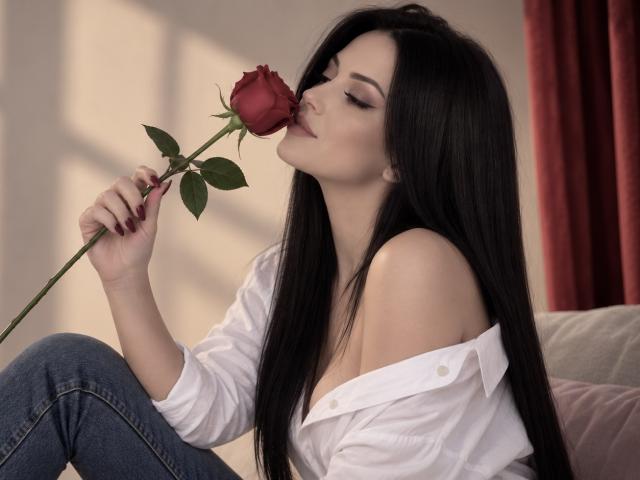 Coryna, com longos cabelos pretos, beijando uma rosa vermelha, vestindo uma camisa branca com um ombro descoberto e jeans azuis, sentada em um sofá, segurando a rosa com folhas verdes..
