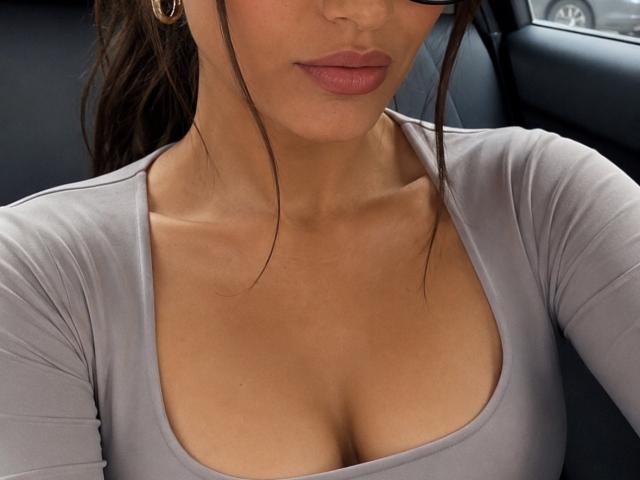 Les seins de Sloaneswan sont visibles dans un haut gris clair moulant avec un décolleté bas, alors qu'elle est assise dans une voiture. Elle a une peau bronzée, des lèvres pulpeuses et des cheveux bruns attachés en queue de cheval..