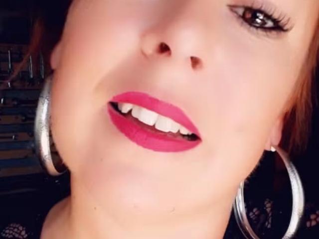 LaFrancaiseGwendoline's face, featuring pink lipstick, white teeth, black eyeliner, and silver hoop earrings, with a slight upward tilt. Her skin is fair, and she's looking at the camera.