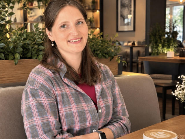 LoraParkerHot, with brown hair and a pink checkered shirt, smiling at the camera, sitting in a cafe, wearing a red top and a black watch.