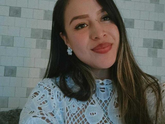LiaDiane, con su largo cabello castaño, lleva una blusa blanca de encaje, tiene unos pendientes bonitos y una ligera sonrisa en su lápiz labial rojo, mostrando su rostro y busto suaves y sensuales..
