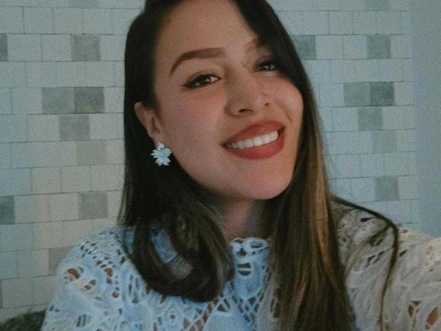LiaDiane with long brown hair, wearing white lace top, smiling, showing teeth, wearing white flower earrings, has light skin, and brown eyebrows, looking cute.