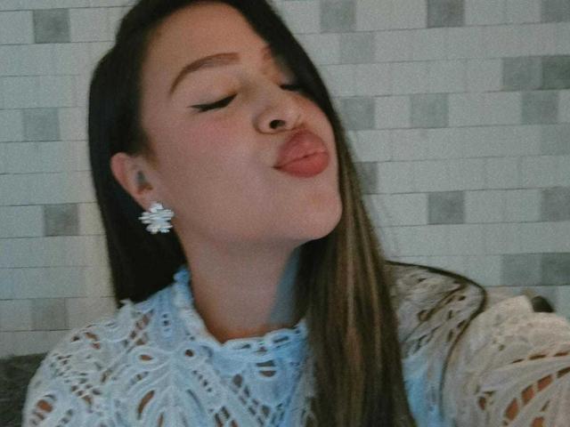 LiaDiane, con cabello castaño largo, ojos cerrados, labios rosados y fruncidos, vistiendo una blusa blanca de encaje, aretes de flores y un piercing en la nariz, luciendo linda y sexy..