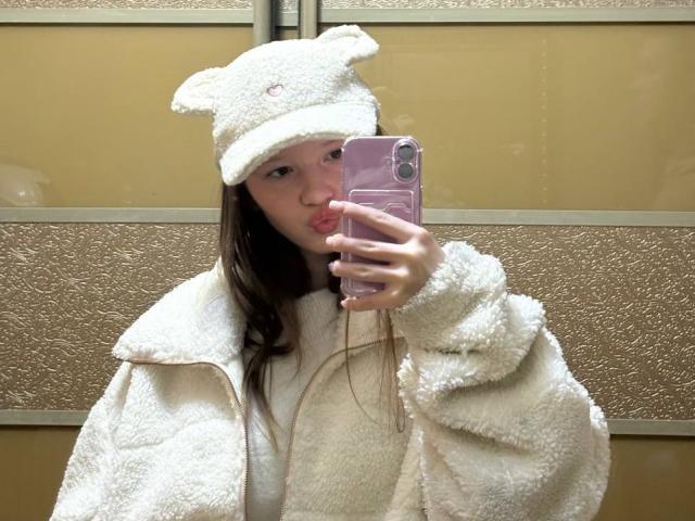 LanieDistel with pouty lips, holding pink phone, wearing fluffy white coat and bear hat, taking mirror selfie, brown hair, fair skin, in front of yellow textured wall.