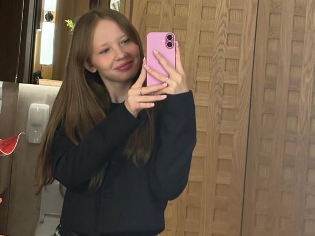 LanieDistel, con lunghi capelli castani, sorridente, che tiene in mano un telefono rosa, indossa un maglione nero, sta scattando un selfie allo specchio in bagno, con un armadietto di legno sullo sfondo..