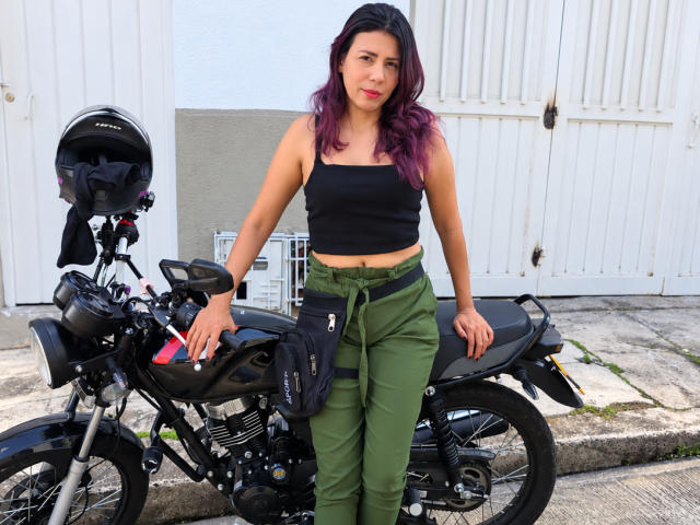 LunaTomson met paars gekleurd haar, een zwart tanktop, groene broek, staat bij een zwarte motorfiets met een helm op het stuur, handen op de motor..