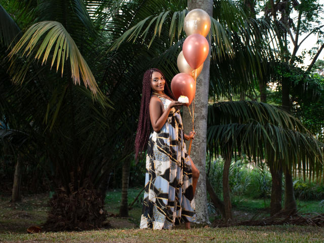 CandiNahomi, con lunghi capelli rossi intrecciati, tiene in mano palloncini rosa e dorati, indossa un vestito a fantasia e si trova in un'area erbosa con palme. Sorride ed è molto carina..