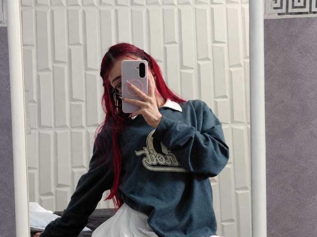 CherryBuchel with red hair, wearing a green hoodie with a logo, white shirt, and black mask, taking a mirror selfie, showing her face partially hidden by her phone.