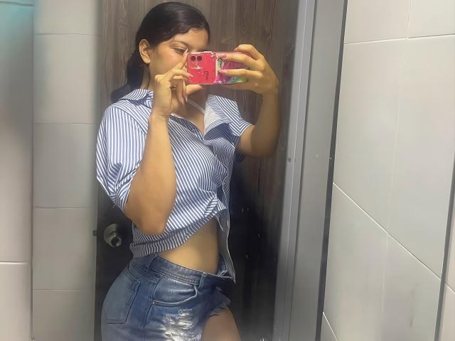 Victoria Fiore, vêtue d'une chemise bleue à rayures nouée, laissant apparaître son ventre, et de shorts en jean déchirés, prend un selfie devant un miroir avec un téléphone rose, montrant ses fesses et ses seins de taille moyenne..