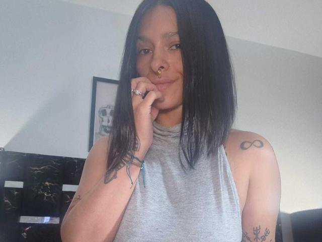 AmelieBlake has black shoulder-length hair, a nose ring, and tattoos on her arm and shoulder. She's wearing a grey sleeveless top, smiling, and touching her chin.