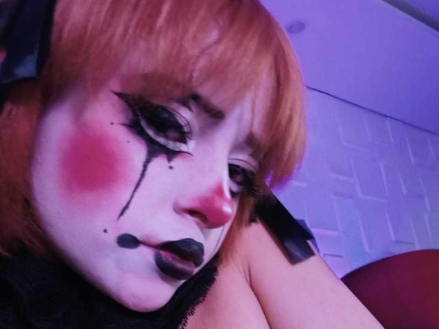 ViolettClay with painted white face, black eyeliner, pink blush, black lipstick, and tears, has red hair, black headband, and is looking down with a sad expression, showing her tits.