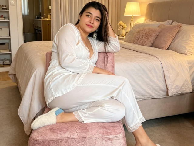 AnyClaus with dark hair, white satin pajamas, and white fluffy slippers, sitting on a pink velvet chair, her small ass and tits visible, in a cozy bedroom.