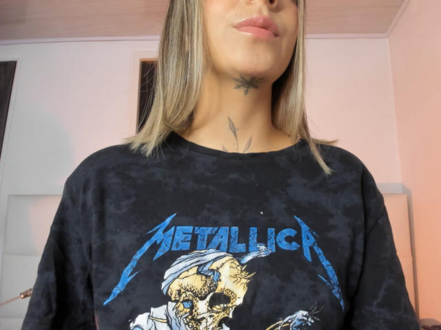 KathyM with blonde hair, wearing a black Metallica t-shirt, has a tattoo on her neck and lower chin, and pinkish lipstick, standing in a room.