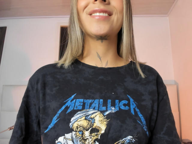 KathyM with straight blonde hair, smiling, wearing a black Metallia shirt, showing a skull and blue text. Visible tattoos on neck and throat.