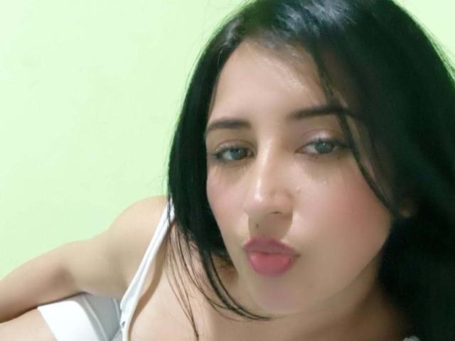 NicollJeans has black hair, blue eyes, and pink lips, pouting slightly with a shiny, sweaty face, wearing a white top, showing her small, perky tits.