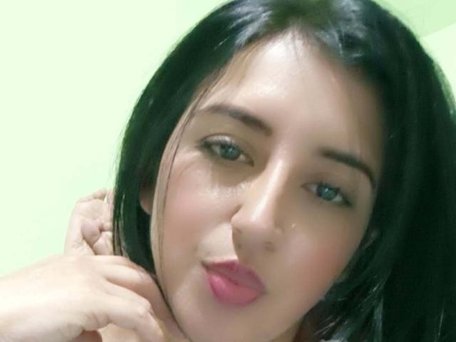 NicollJeans with black hair, blue eyes, and pink lipstick, pouting slightly, touching her neck with one hand. Her skin is fair, and she has a subtle smile.