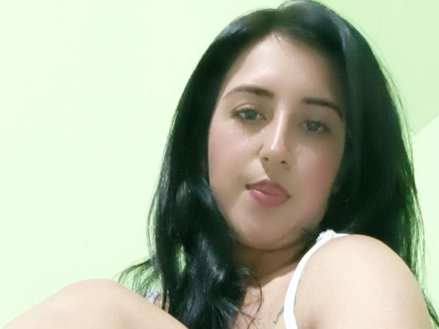 NicollJeans has black hair, light skin, and is looking at the camera with a slight smile, wearing a white top, showing her bare shoulders and upper chest.