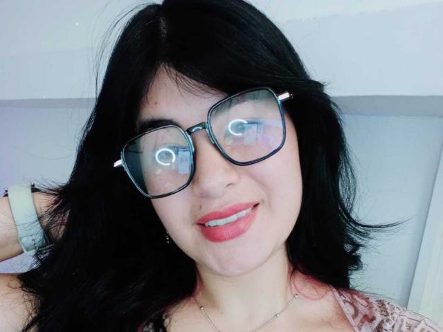 NinaHarpper, com cabelo preto, óculos e batom rosa, mostrando o reflexo nos óculos, sorrindo com os lábios vermelhos ligeiramente entreabertos, vestindo uma blusa de renda rosa e uma pulseira branca..