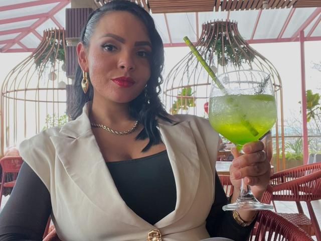 JessicaFiore, con capelli scuri, rossetto rosso, orecchini dorati e una giacca bianca, tiene in mano un cocktail verde con una cannuccia, indossa una maglietta nera, una collana dorata ed è seduta a un tavolo di vimini rosso, guardando verso la fotocamera..