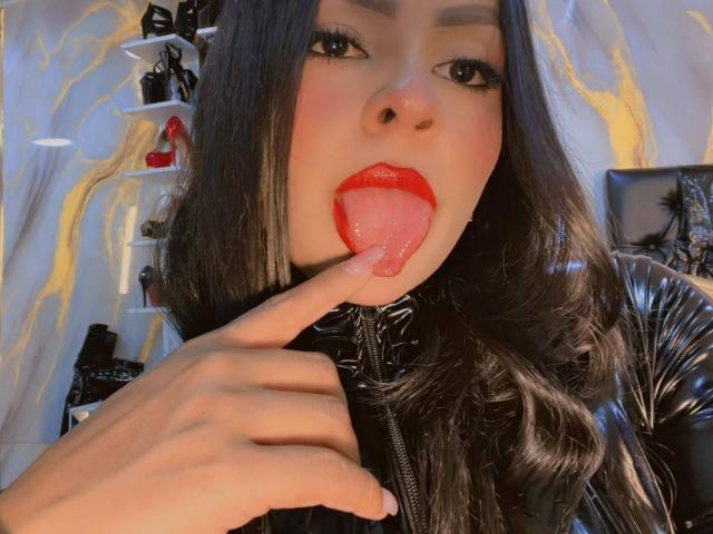 LindaFrancoo, con cabello negro largo, lápiz labial rojo y una chaqueta de cuero negro, saca la lengua y la toca con su dedo. Sus senos son visibles..