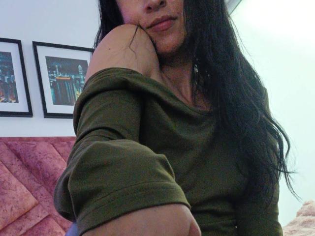ZarayFeet with long black hair, green off-shoulder top, and bare shoulder, resting chin on arm, pink couch, and framed pictures in the background. Her tits are partially visible.