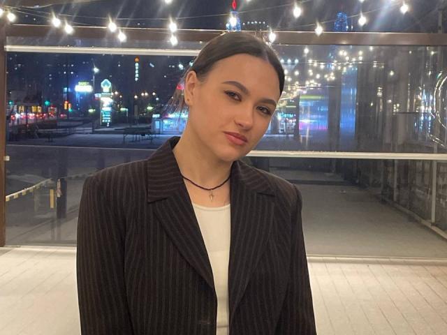 Isobel Staehle, com tranças escuras, um blazer preto listrado, uma camisa branca, maquiagem discreta e um colar preto, sentada em uma área bem iluminada, com luzes de Natal, à noite..