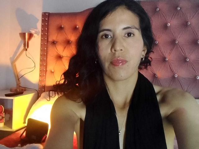 LarisaBrown with dark wavy hair, black scarf, and shiny pink lips, sitting in bed, head against tufted pink headboard, with a gold lamp on the left.
