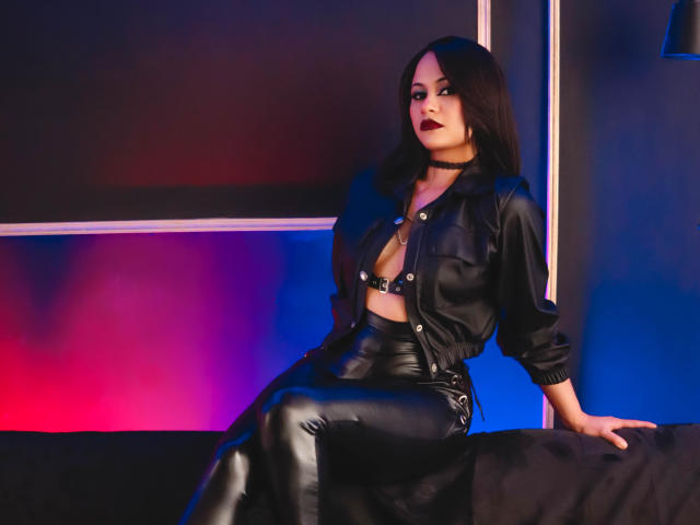 CassieJones with black leather pants, open black jacket, black choker, and dark lipstick, sitting on a black couch, showcasing her small tits and shiny black leather booty.