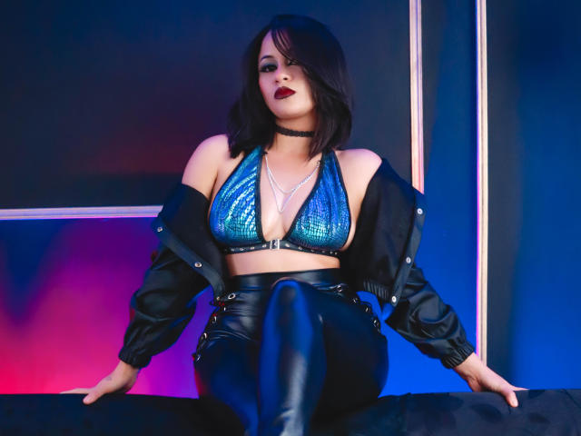 Cassie Jones, avec des seins ornés de paillettes bleues, un pantalon en cuir noir et une veste noire, assise les jambes jointes, portant un collier noir, dans un décor éclairé de bleu et rose néon..