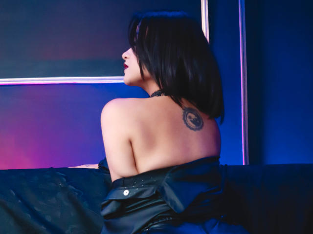 Cassie Jones, avec ses cheveux noirs, son dos dégagé, une veste en cuir noir et un petit tatouage sur le haut du dos, assise avec la tête tournée sur le côté, a l'air séduisante..