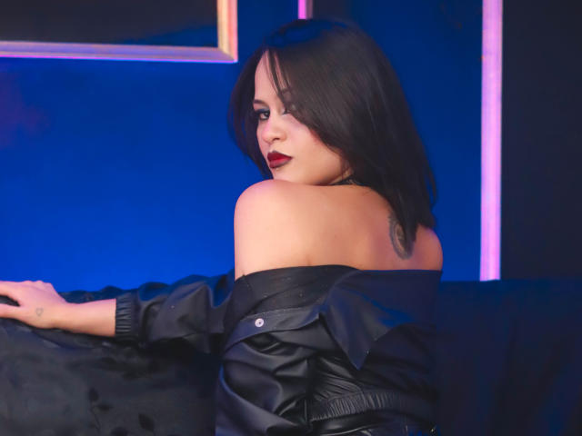CassieJones with dark hair, red lipstick, and black off-shoulder leather jacket, showing bare shoulder, looking over shoulder, sitting on black couch, blue neon light in background.