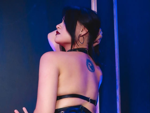 Cassie Jones, avec ses cheveux noirs, son rouge à lèvres foncé et ses boucles d'oreilles, porte un haut en cuir noir avec des bretelles qui expose son dos avec un tatouage, et pose près d'un poteau..