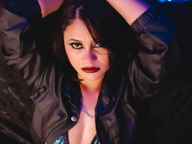 Cassie Jones, avec ses cheveux foncés, sa veste en cuir noir et son rouge à lèvres rouge, les bras levés, regardant vers le haut, avec un léger clin d'œil, portant un collier en argent, et affichant une expression sexy et confiante..